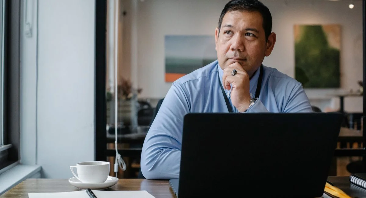 Concentrated ethnic adult male entrepreneur thinking on project while working at table with netbook and cup of drink